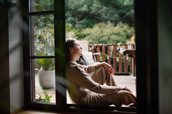 Woman resting in doorway of balcony doors, resting. Single woman enjoying free weekend. Concept of selfcare. Woman using smart phone